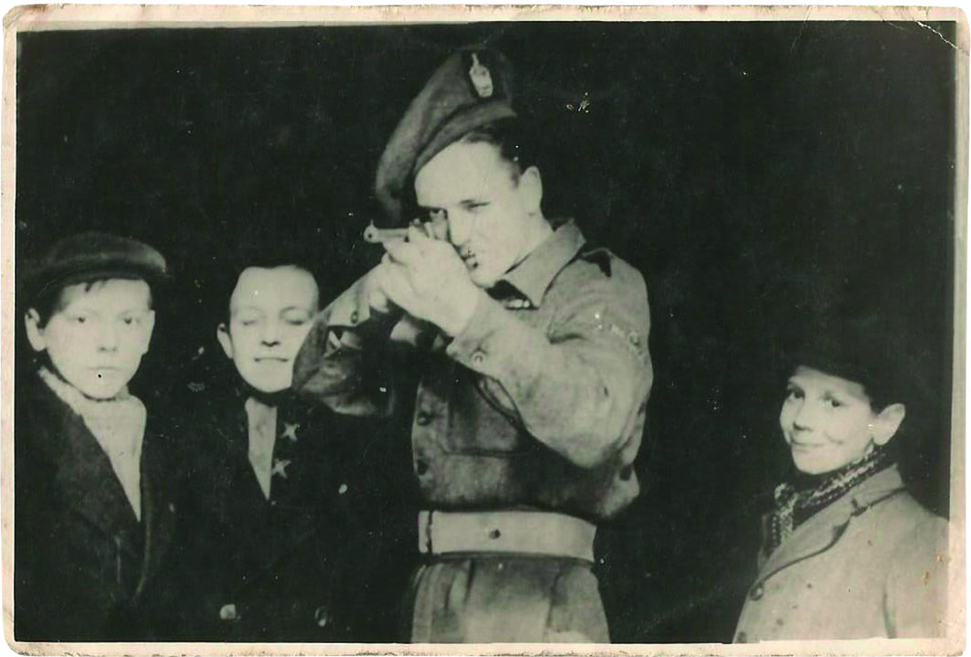 Black and white photograph of a man in Second World War army uniform shooting a rifle while three little boys look on