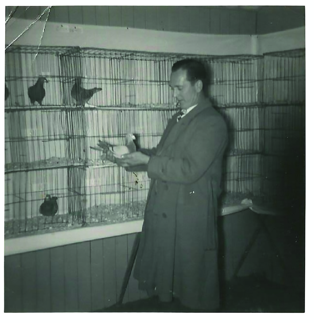 Black and white photograph of a man in a trench coat standing in front of some cages of pigeons