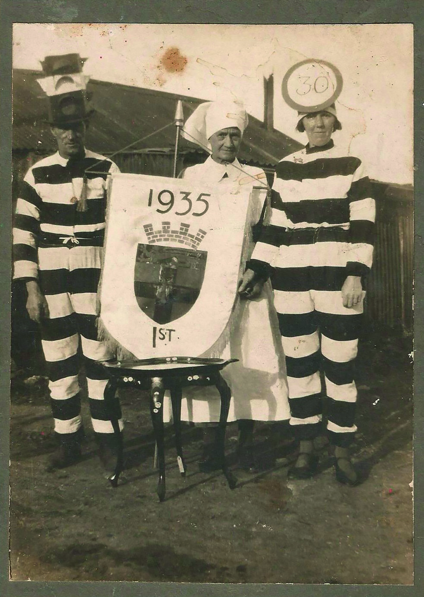 Black and white photograph of a man wearing a black-and-white striped outfit and a hat made to look like a set of traffic lights, an older woman in a nurse's costume holding a shield that says '1935 - 1st' and a woman in a black-and-white striped outfit with a '30' speed sign on her head