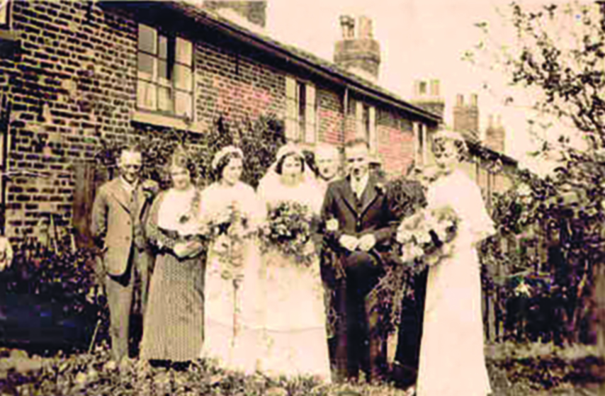 Black and white photograph of a bride posing outside a cottage with her bridesmaids and others.