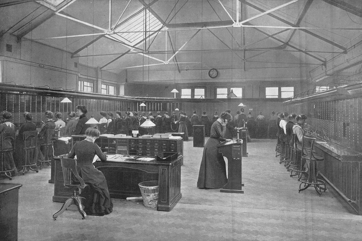 Black and white photograph of a large room with a row of women in Edwardian dress sitting on either side operating switchboards, while more women take telephone calls in the middle
