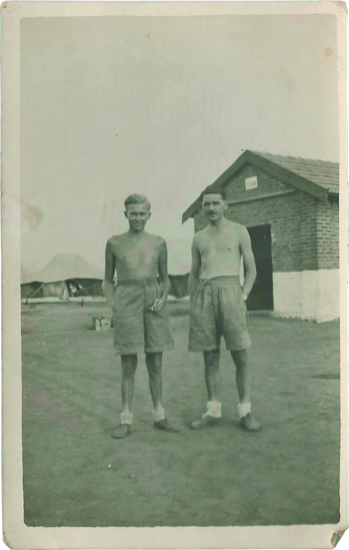 Black and white photograph of two young men wearing shorts and no shirts standing in a military base
