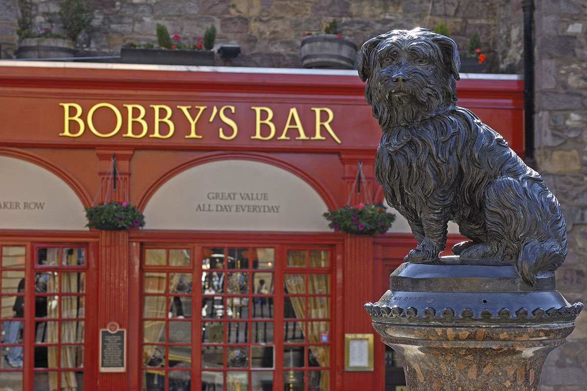 A metal statue of Greyfriars Bobby, a Skye terrier, in front of a bar with a red pub sign saying 'Bobby's Bar' in gold lettering