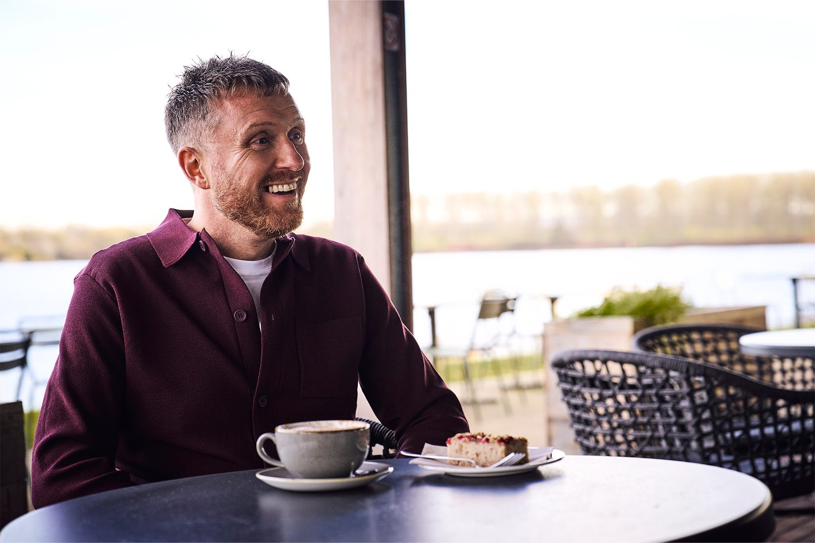 Michael, a man with a gingery bear sits at a cafe table with tea and cake, smiling at someonee out of frame