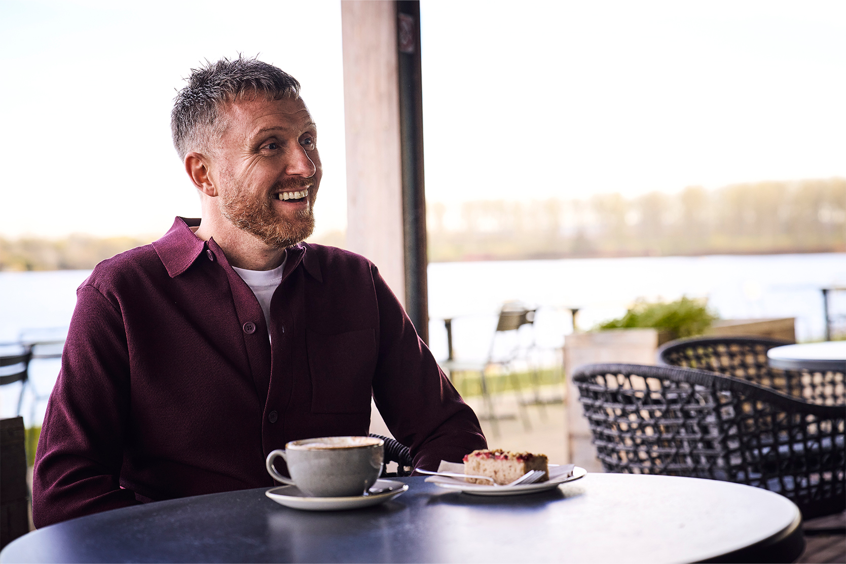 Michael, a man with a gingery bear sits at a cafe table with tea and cake, smiling at someonee out of frame
