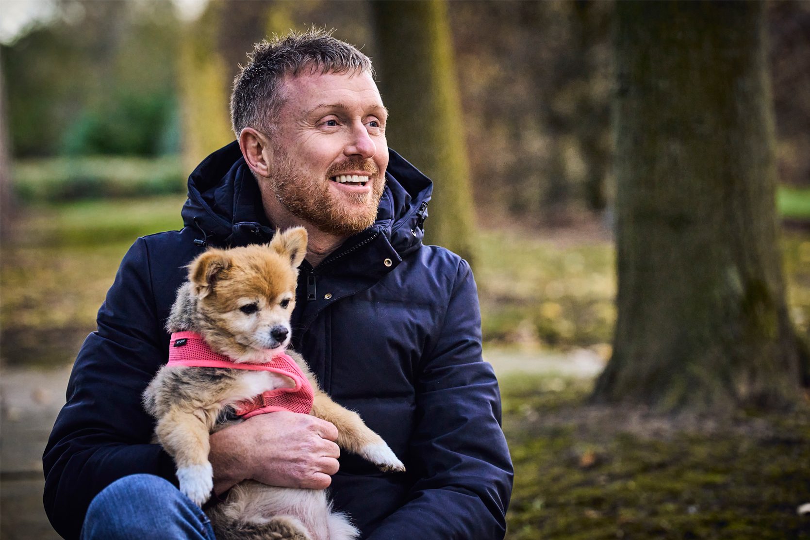 Michael, a man in a winter coat and jeans sits in a park holding his fluffy dog Phyllis