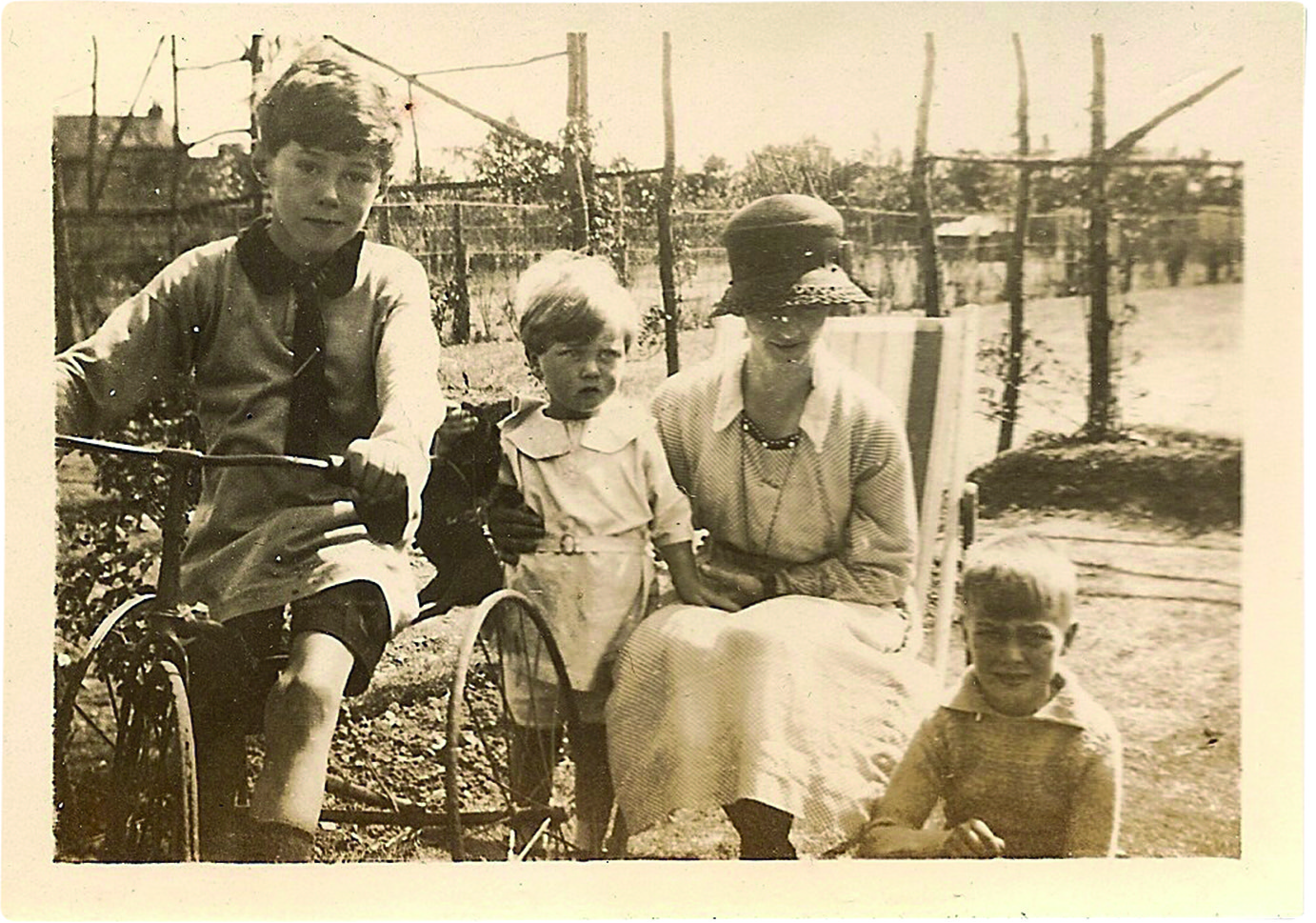 Black and white photograph of a woman in 1920s clothing sitting in a garden with three children, one of whom has a bike
