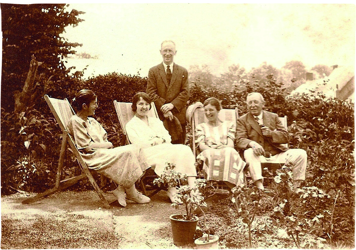 Black and white photograph of three women and a man sitting in deck chairs in a garden while another man stands over them