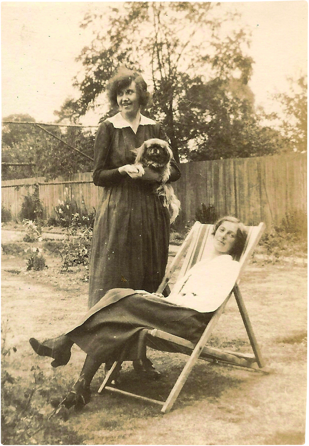 Sepia photograph of a young woman sitting in a deck chair while another woman stands over her holding a Pekinese dog