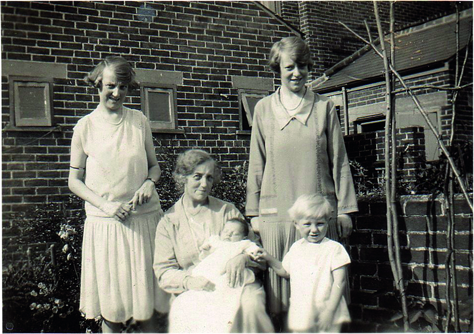 Black and white photograph of two women standing up, an older woman sitting down and holding a baby, and a little boy