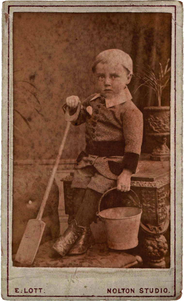 A sepia-tinted old photograph of a little boy in a suit with a bucket and spade